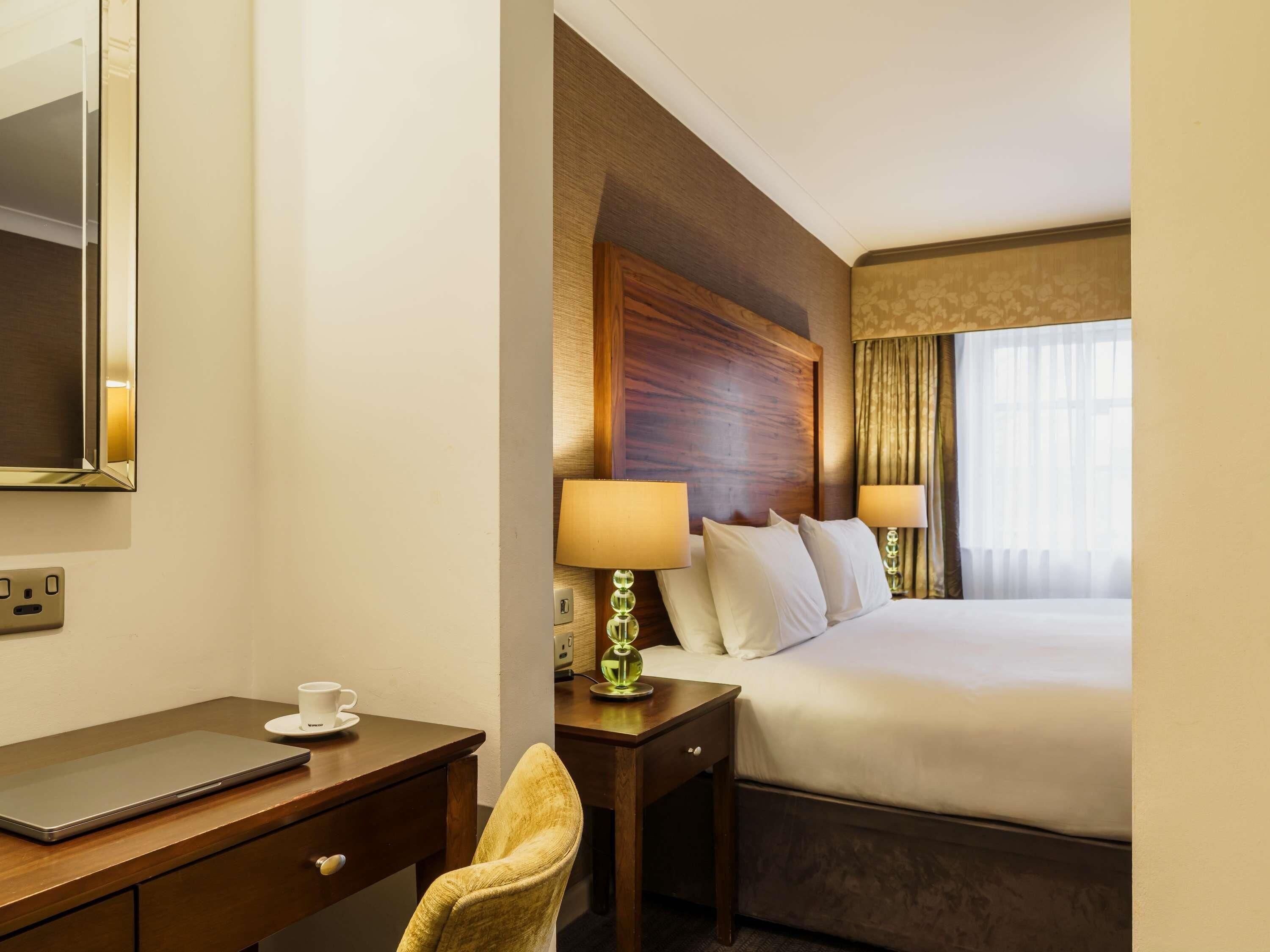 A hotel room with a bed, bedside lamps, a desk with a laptop, a chair, and a mirror.