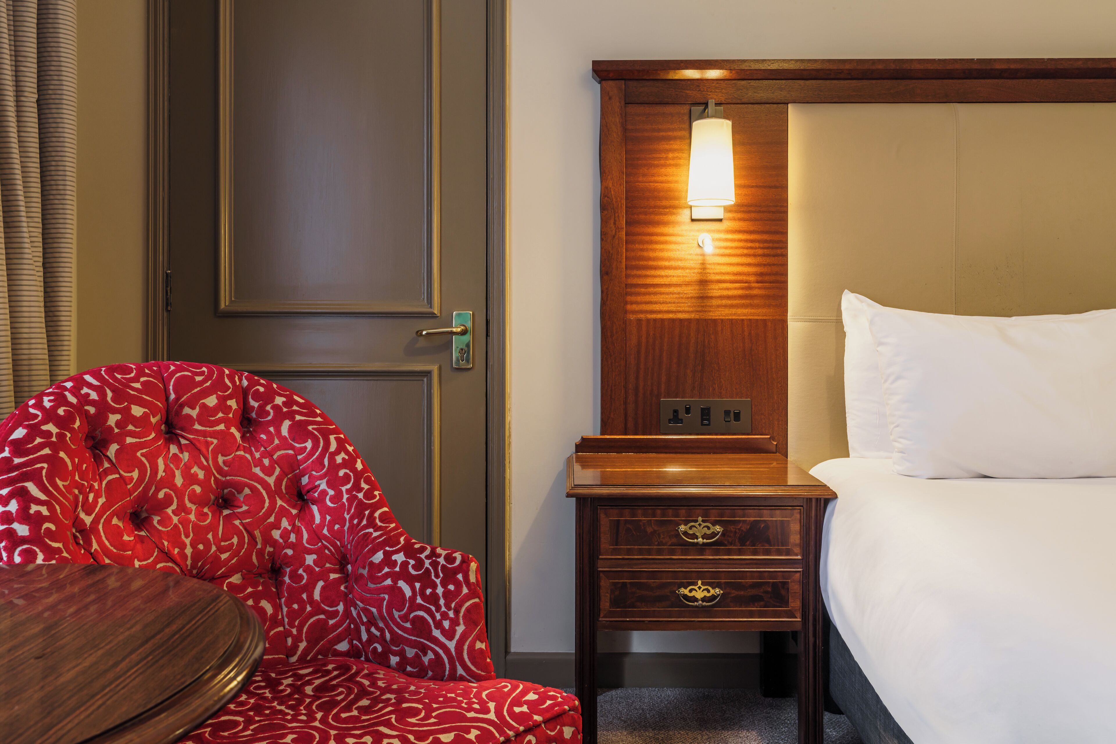 A hotel room with a bed, a bedside table, a lamp, and a red patterned chair.