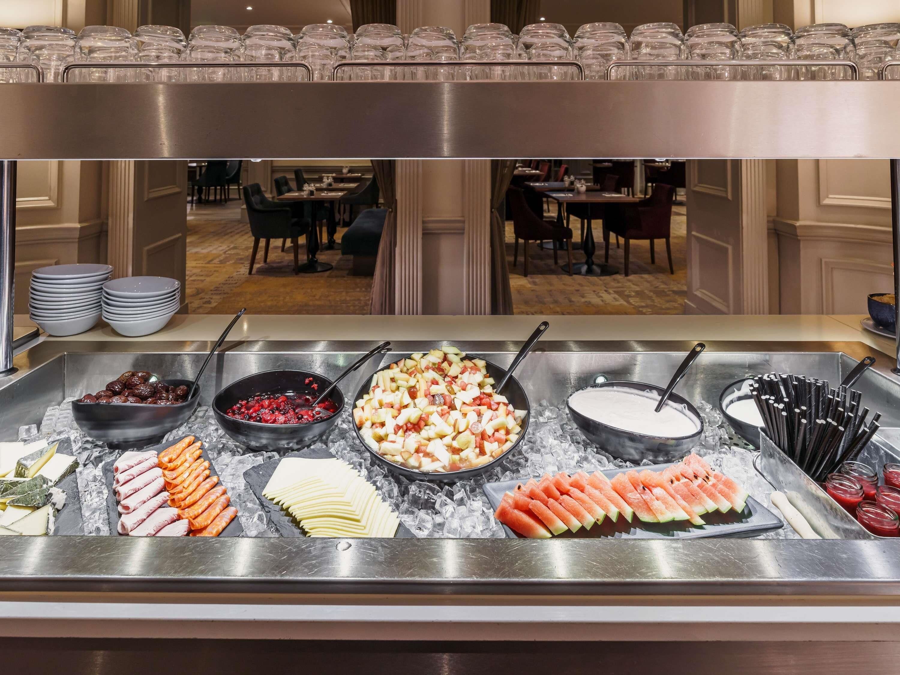 Un buffet proposant une variété de plats, notamment des fruits, des viandes et des desserts, avec la salle à manger en arrière-plan.