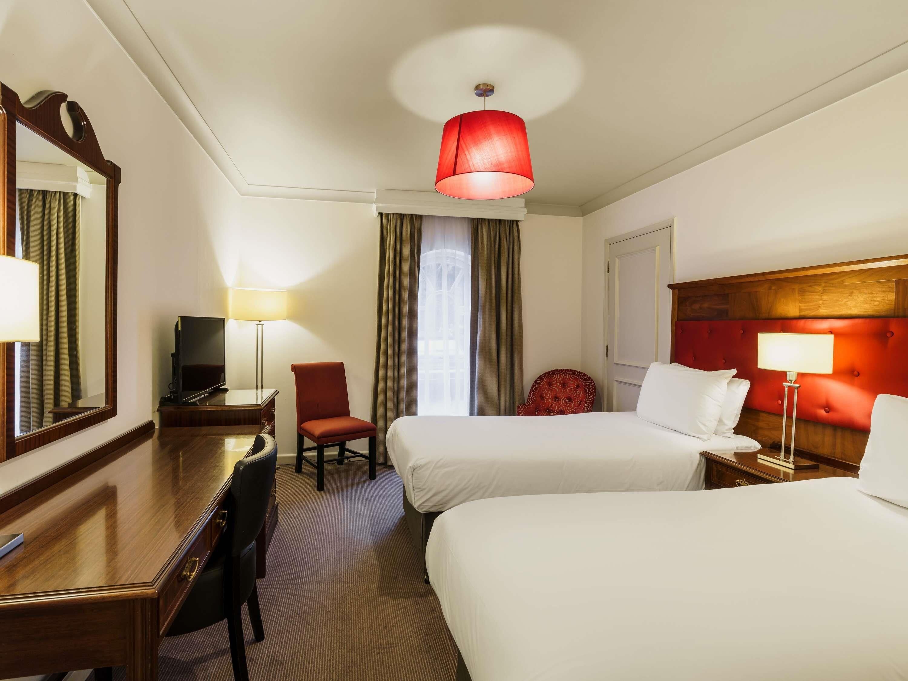 A hotel room with two beds, a desk with a TV, a chair, and a red accent wall.