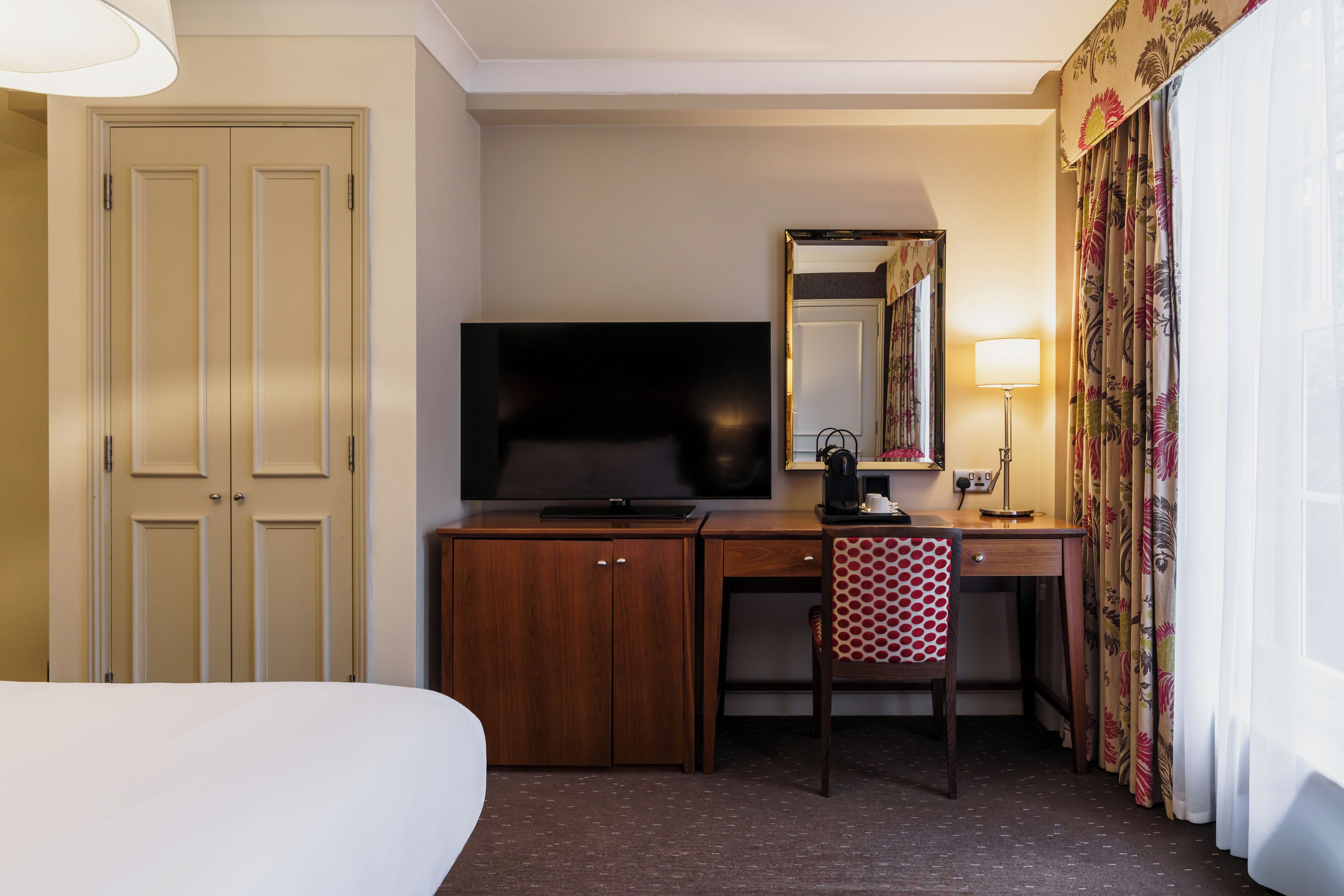 Una camera d'albergo con un letto, una scrivania con televisore a schermo piatto, una sedia, una lampada da tavolo e una finestra con tende.