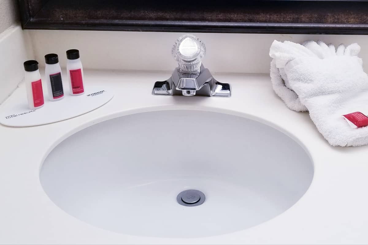 Un lavabo blanc dans une salle de bain, avec un miroir au-dessus, une serviette et trois bouteilles de shampoing.
