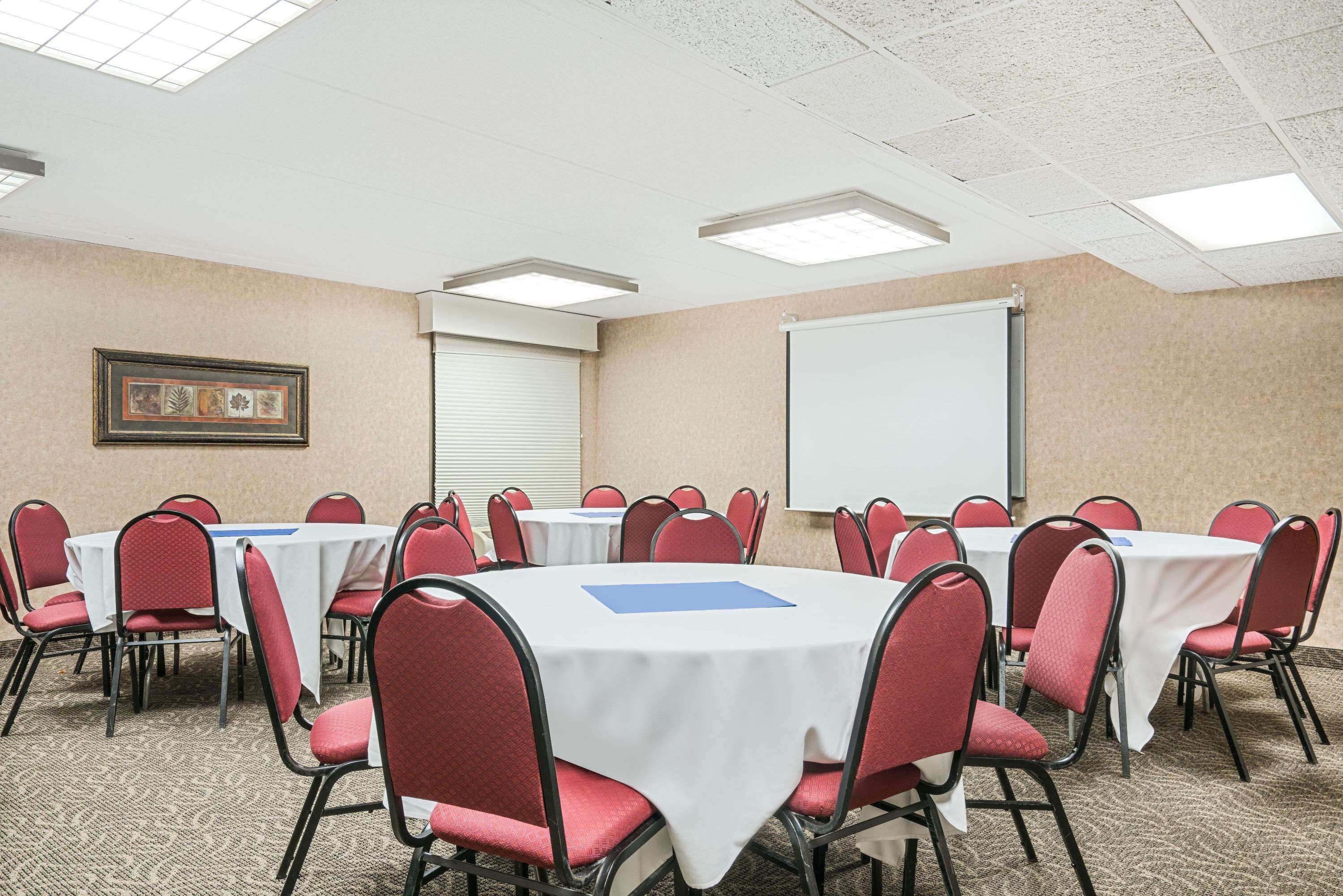 A conference room with round tables, red chairs, white tablecloths, a projector screen, and a framed picture on the wall.