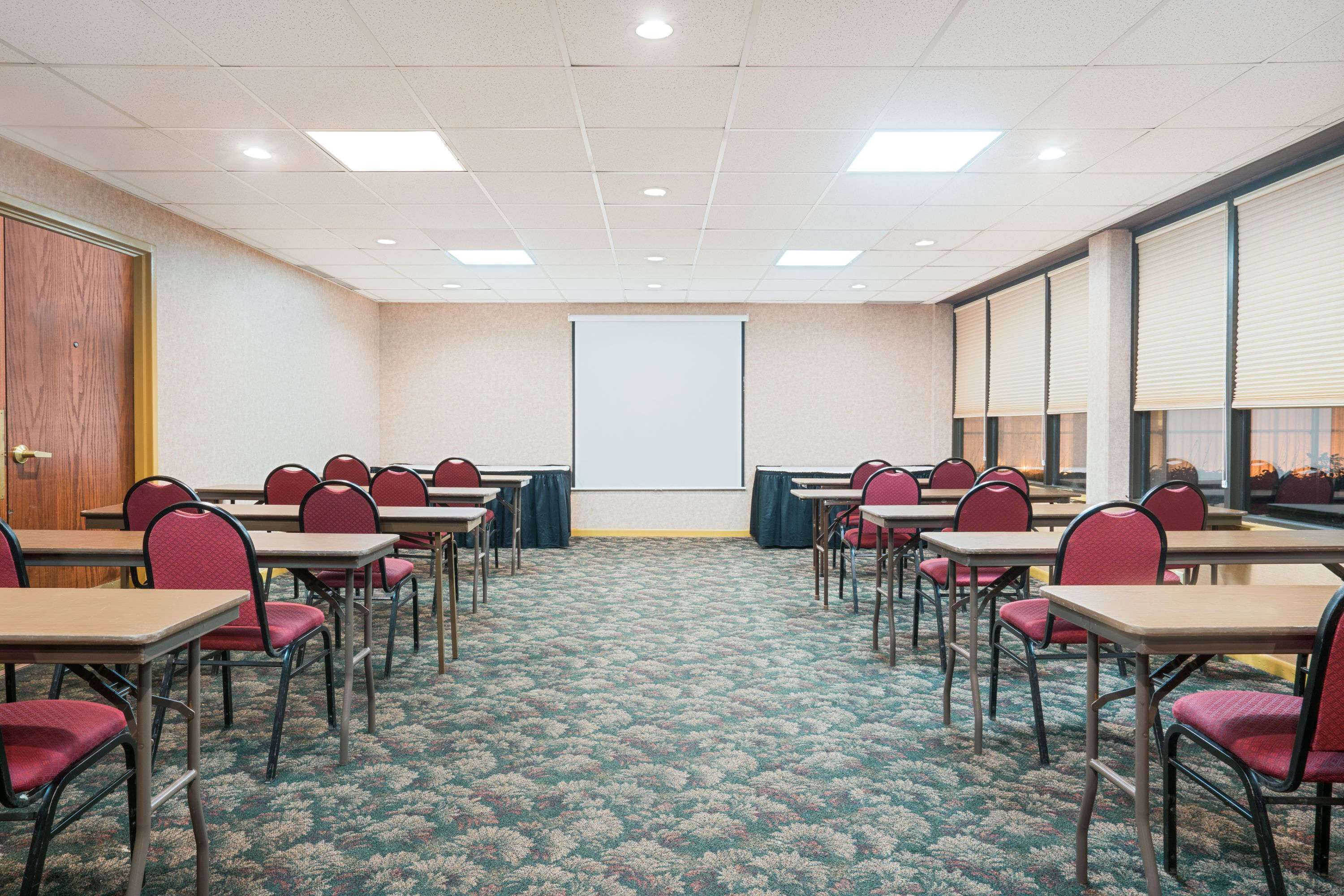 A conference room with tables and chairs arranged for meetings, a projector screen, and a carpeted floor.