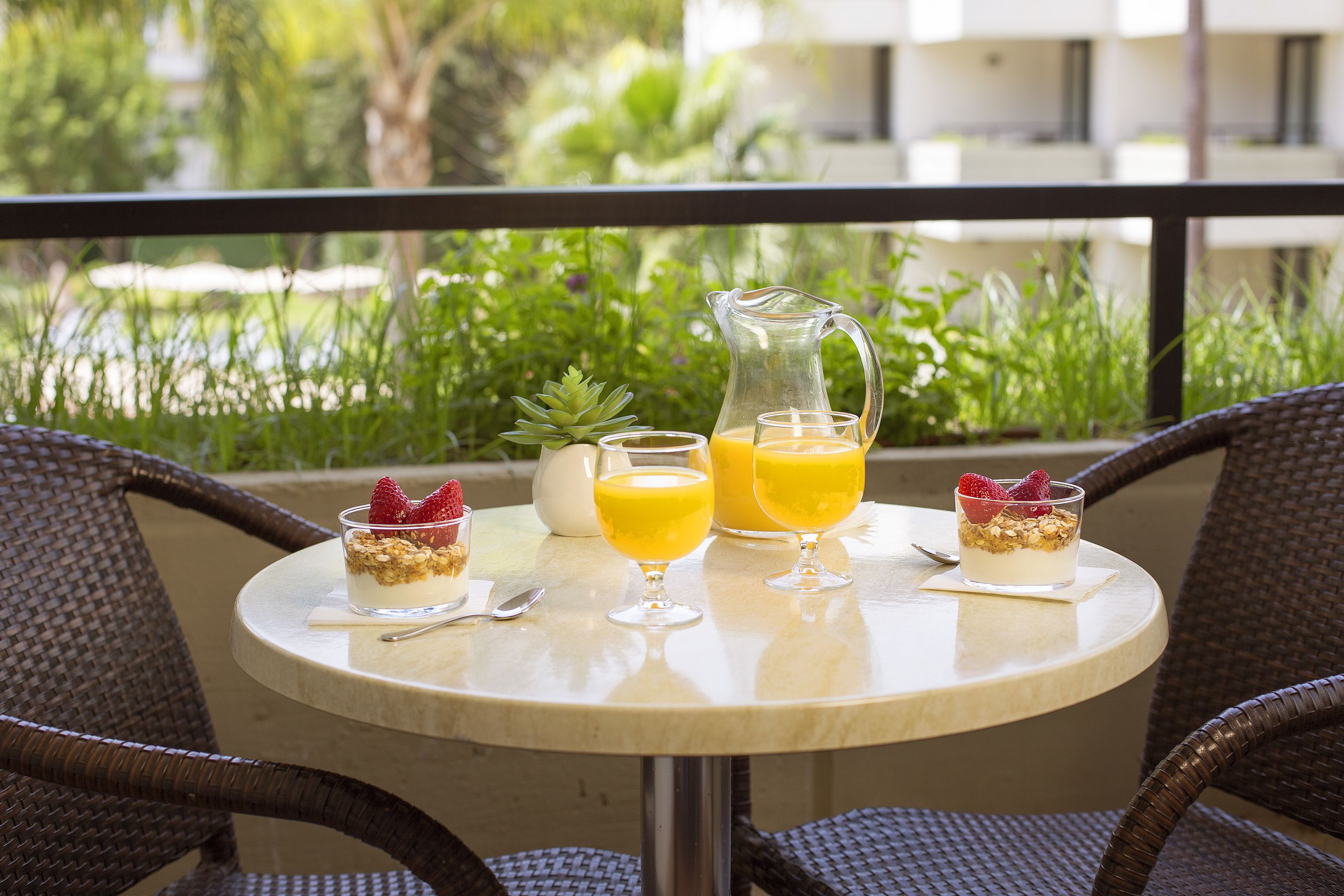 Una mesa redonda con una jarra, dos vasos de jugo de naranja y dos postres con fresas y granola en un balcón con sillas de mimbre.