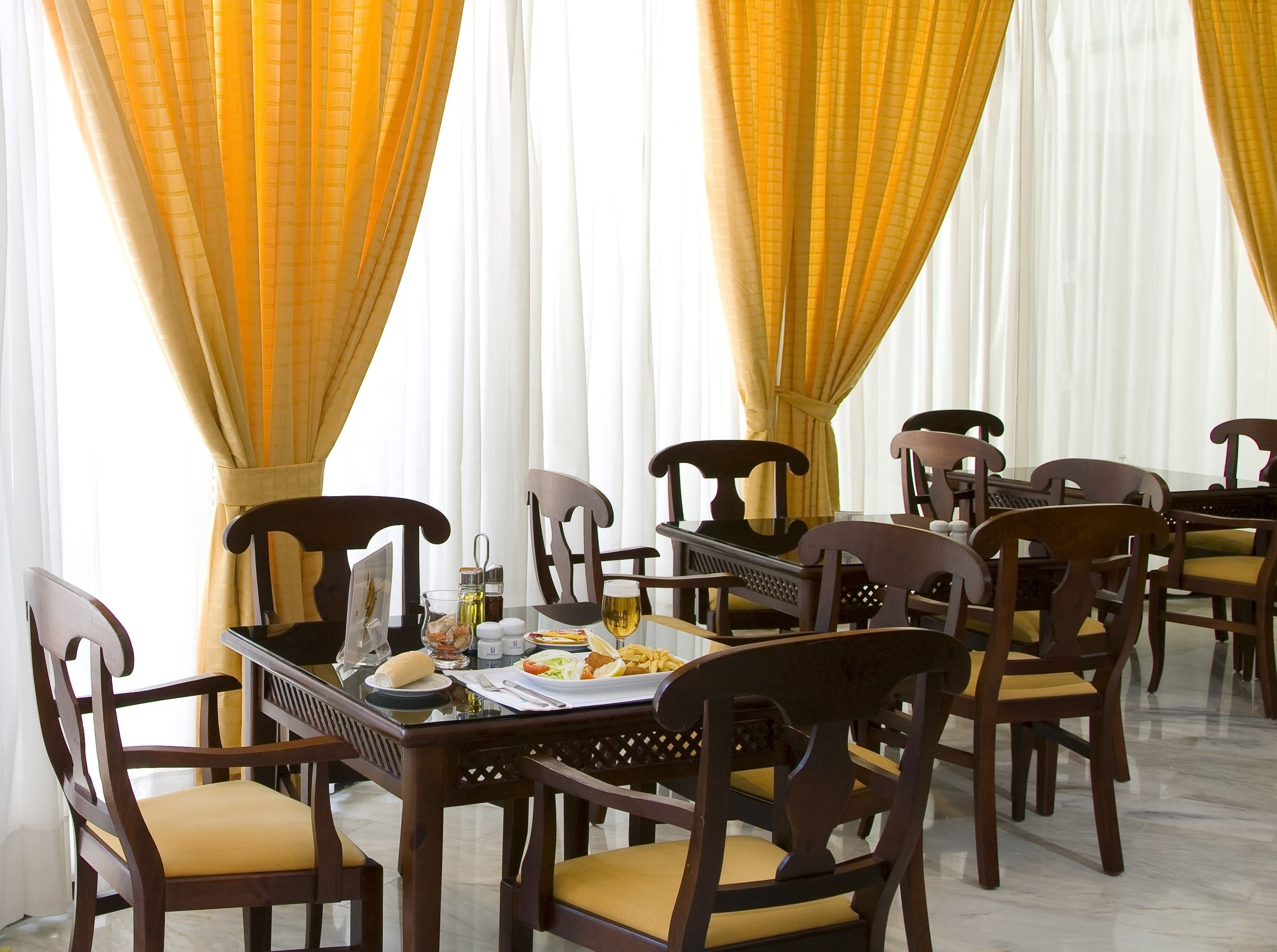 Un coin repas avec des chaises en bois et une table en verre, des rideaux jaunes et une nappe blanche.