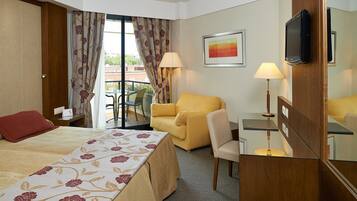A hotel room with a bed, a yellow armchair, a desk, a TV, and a window with curtains.