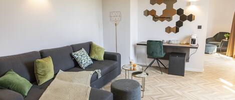 A modern living room with a sofa, ottoman, and coffee table. There is a desk with a chair and a coffee machine. The room has a geometric wall decoration and a floor lamp.