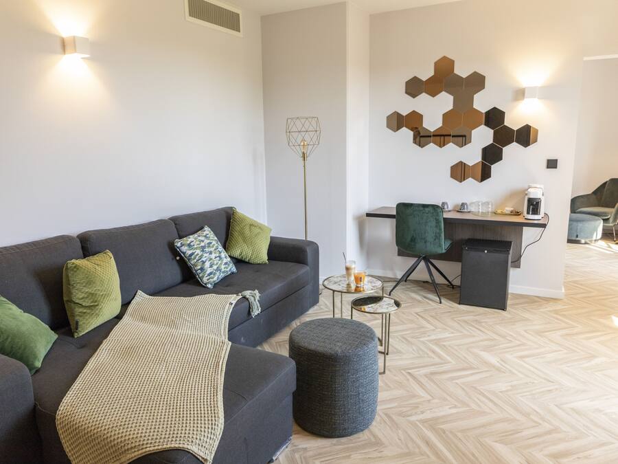 A modern living room with a sofa, ottoman, and coffee table. There is a desk with a chair and a coffee machine. The room has a geometric wall decoration and a floor lamp.