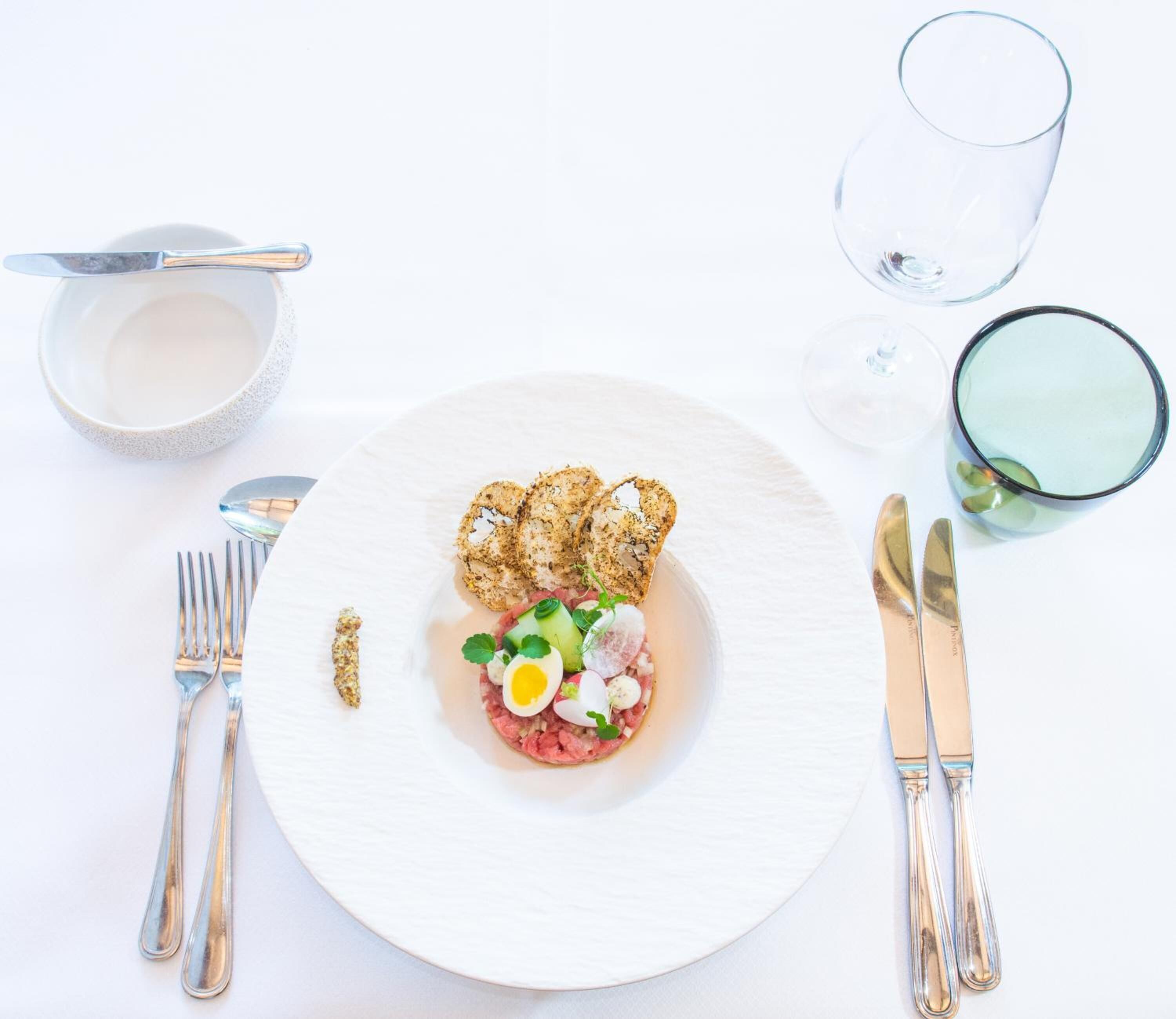 A white plate with a meat-based dish, garnished with herbs and a boiled egg, accompanied by toasted bread and a small bowl of sauce.