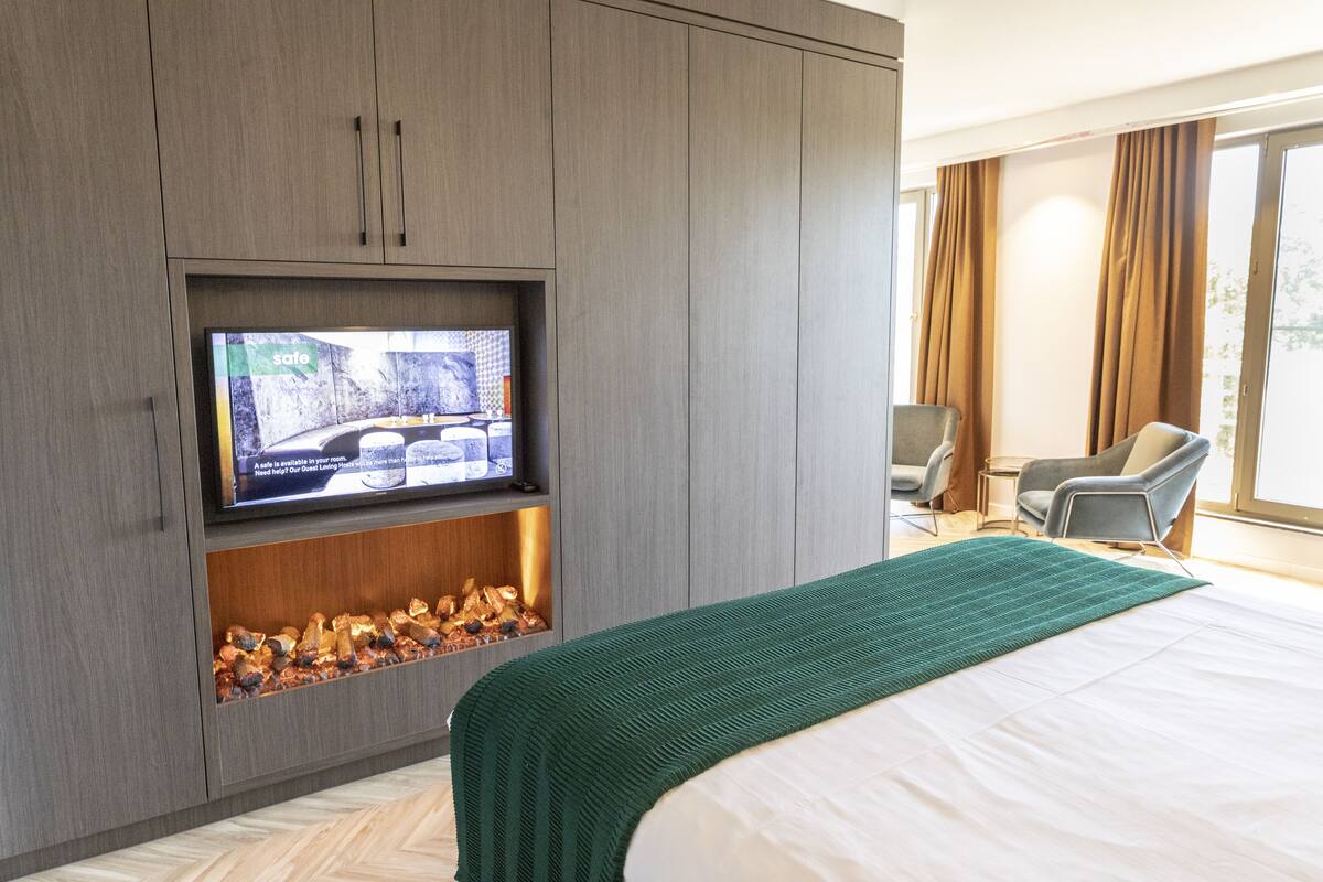 A hotel room with a large bed, a television mounted on a cabinet, a fireplace, and a view of the outdoors.
