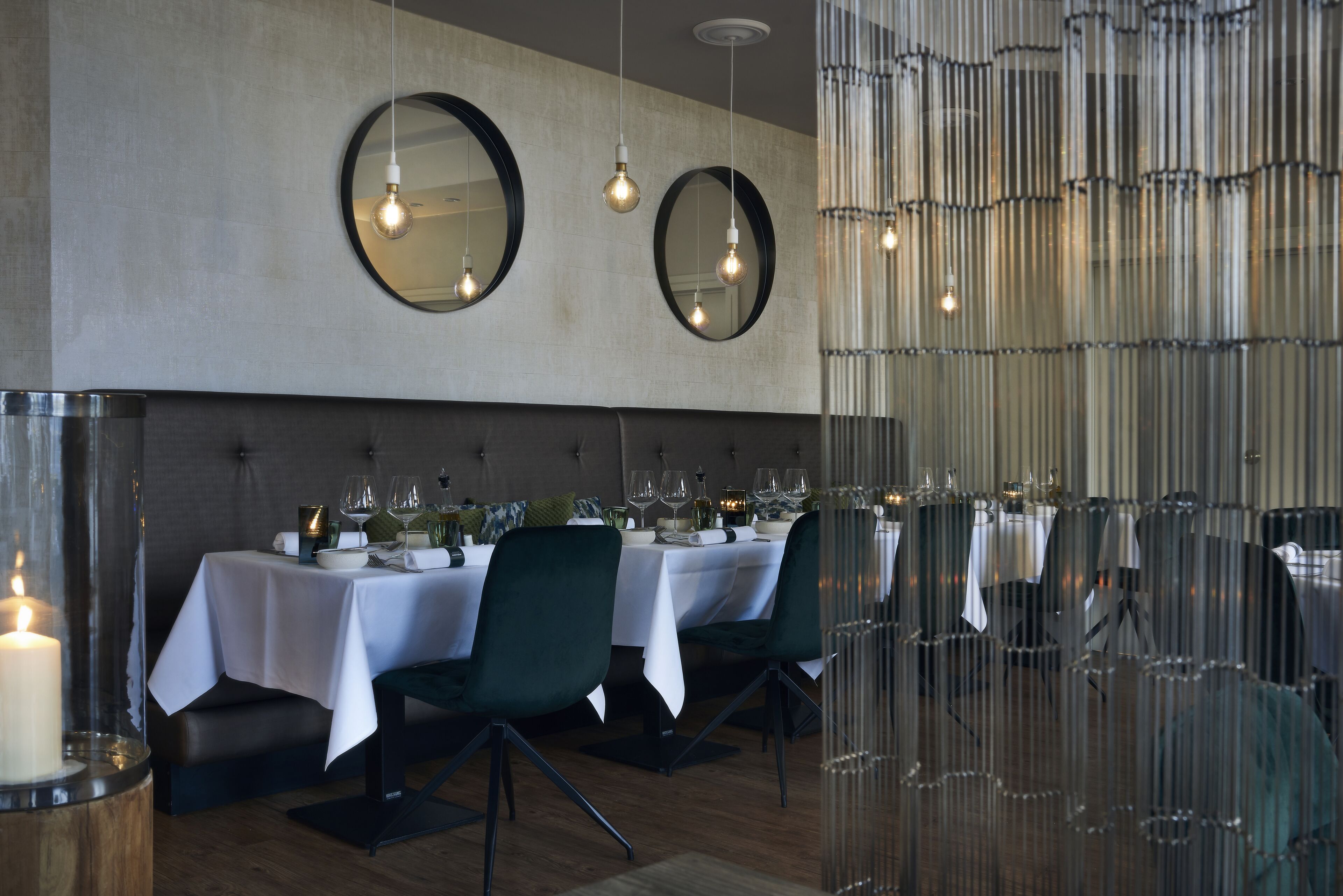 A dining area with round mirrors on the wall, a row of tables with white tablecloths, and a candle in a glass jar.