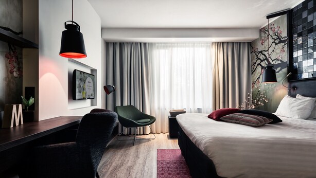 A modern hotel room with a bed, desk, chair, and a large window with curtains.
