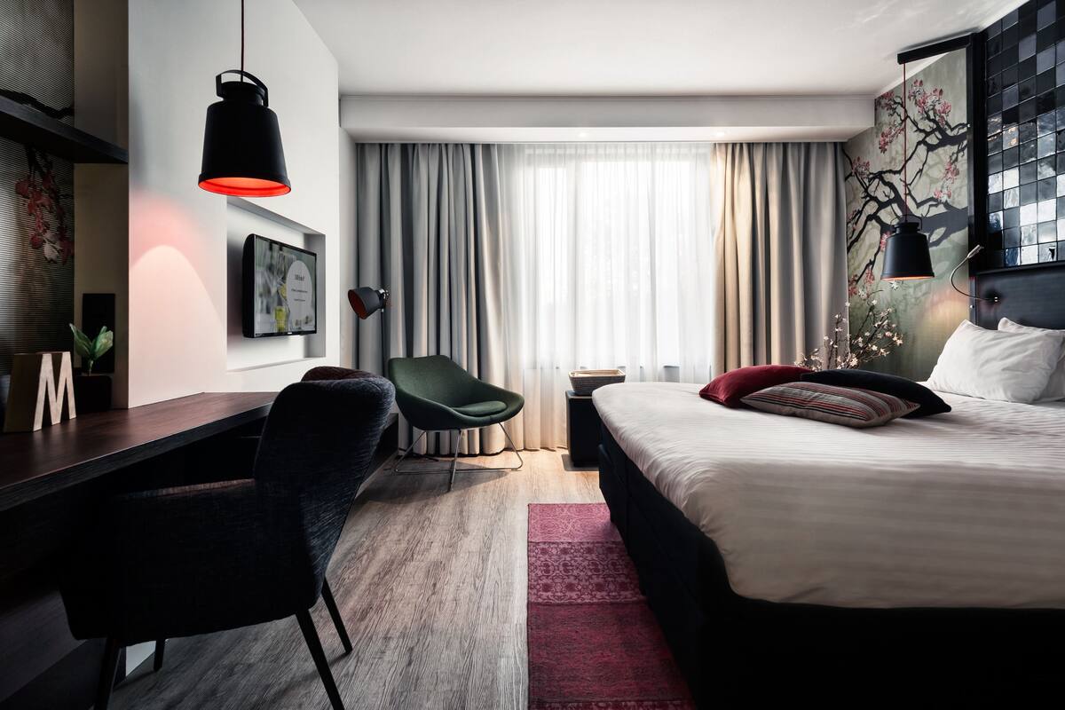 A modern hotel room with a bed, desk, chair, and a large window with curtains.