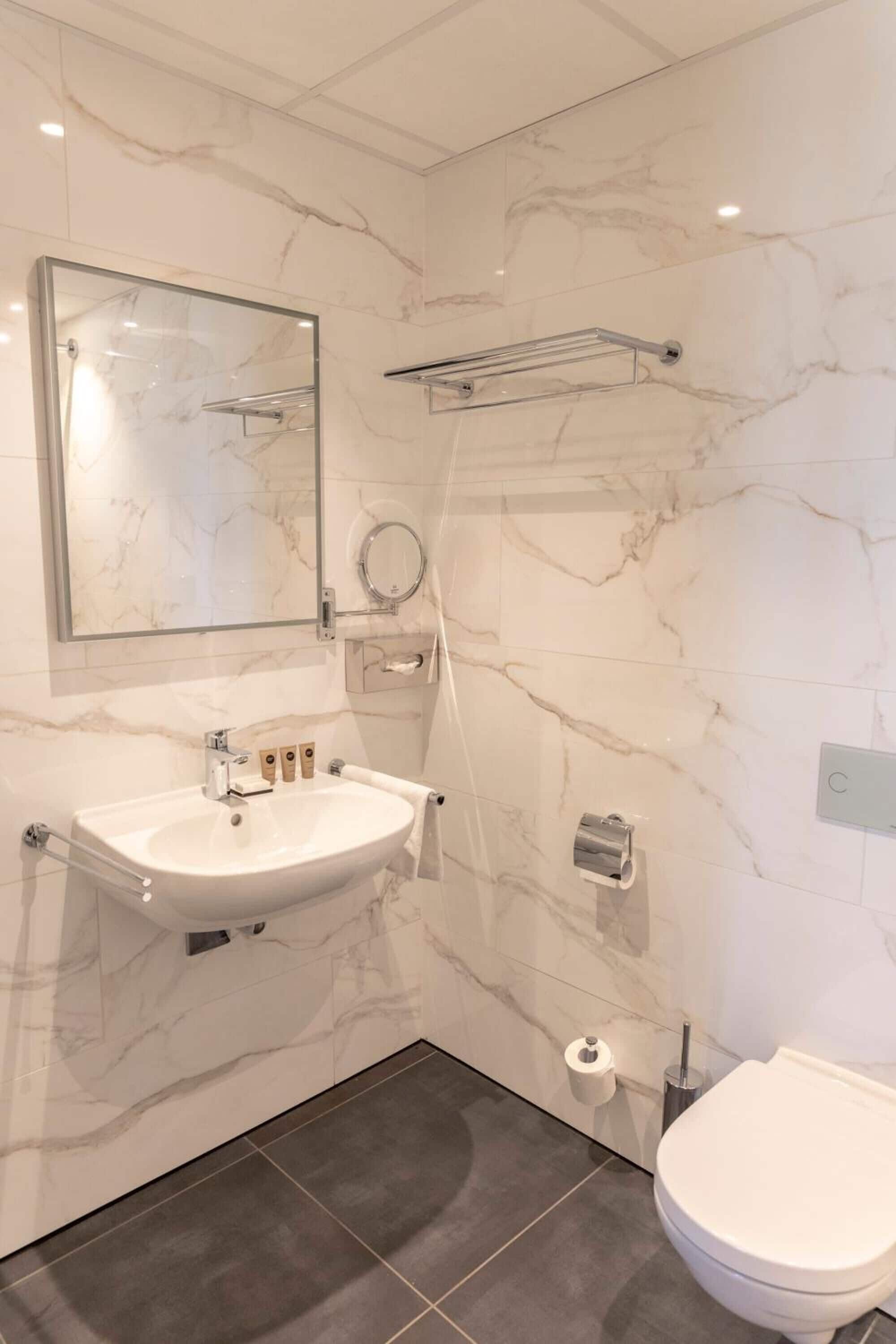 A modern bathroom with a marble wall, a white sink, a mirror, a toilet, and a shower area.