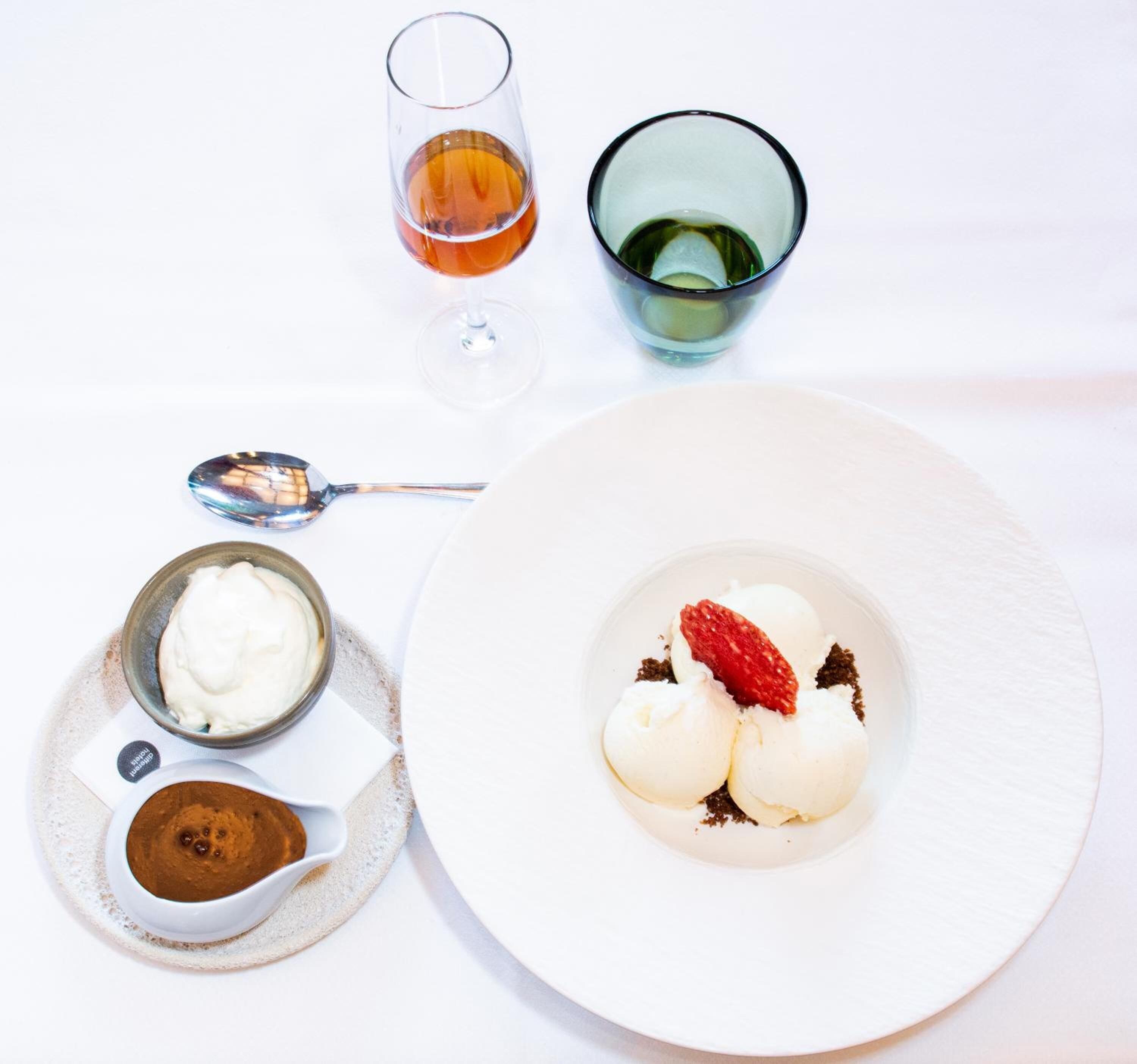 Un plato de postre con helado y una fresa, un vaso de té, un bol de crema batida y una cuchara.