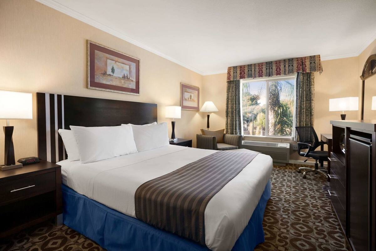 A hotel room with a large bed, two bedside tables with lamps, a desk with a chair, a window with curtains, and a painting on the wall.