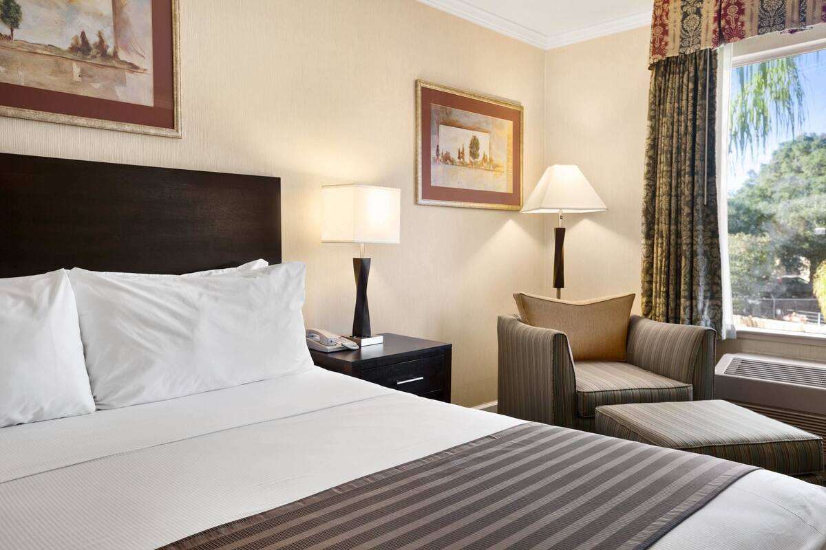 A hotel room with a large bed, two bedside tables with lamps, a chair, and a window with curtains.