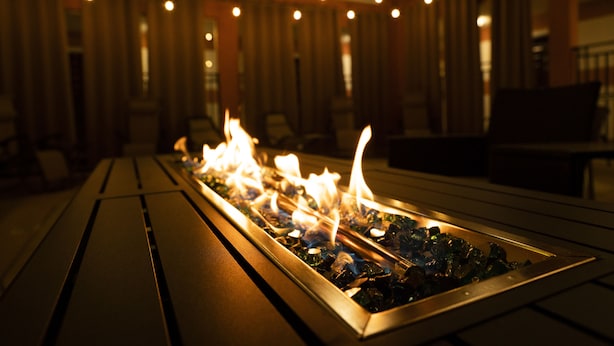 Ein langer, rechteckiger Kamin mit Flammen, in einem Metallrahmen, in einem schwach beleuchteten Innenraum mit Holzvertäfelung und Tischen.