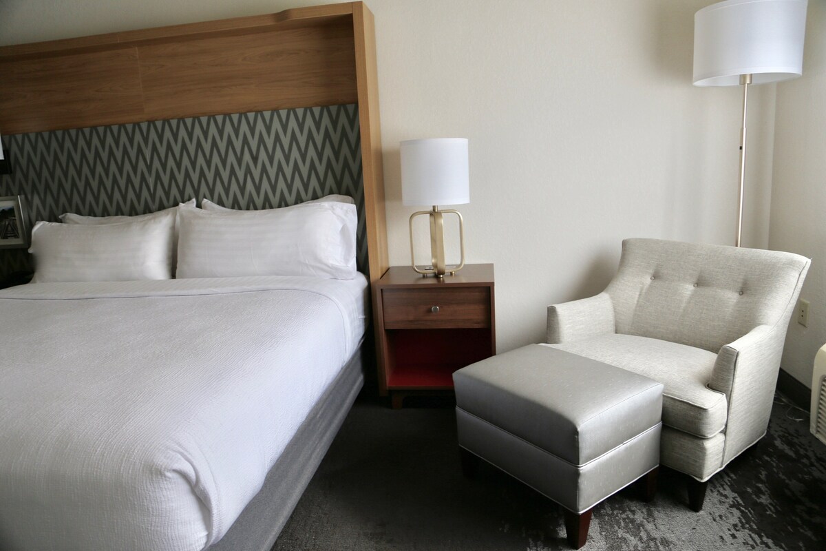 A hotel room with a large bed, a nightstand with a lamp, a chair, and an ottoman.
