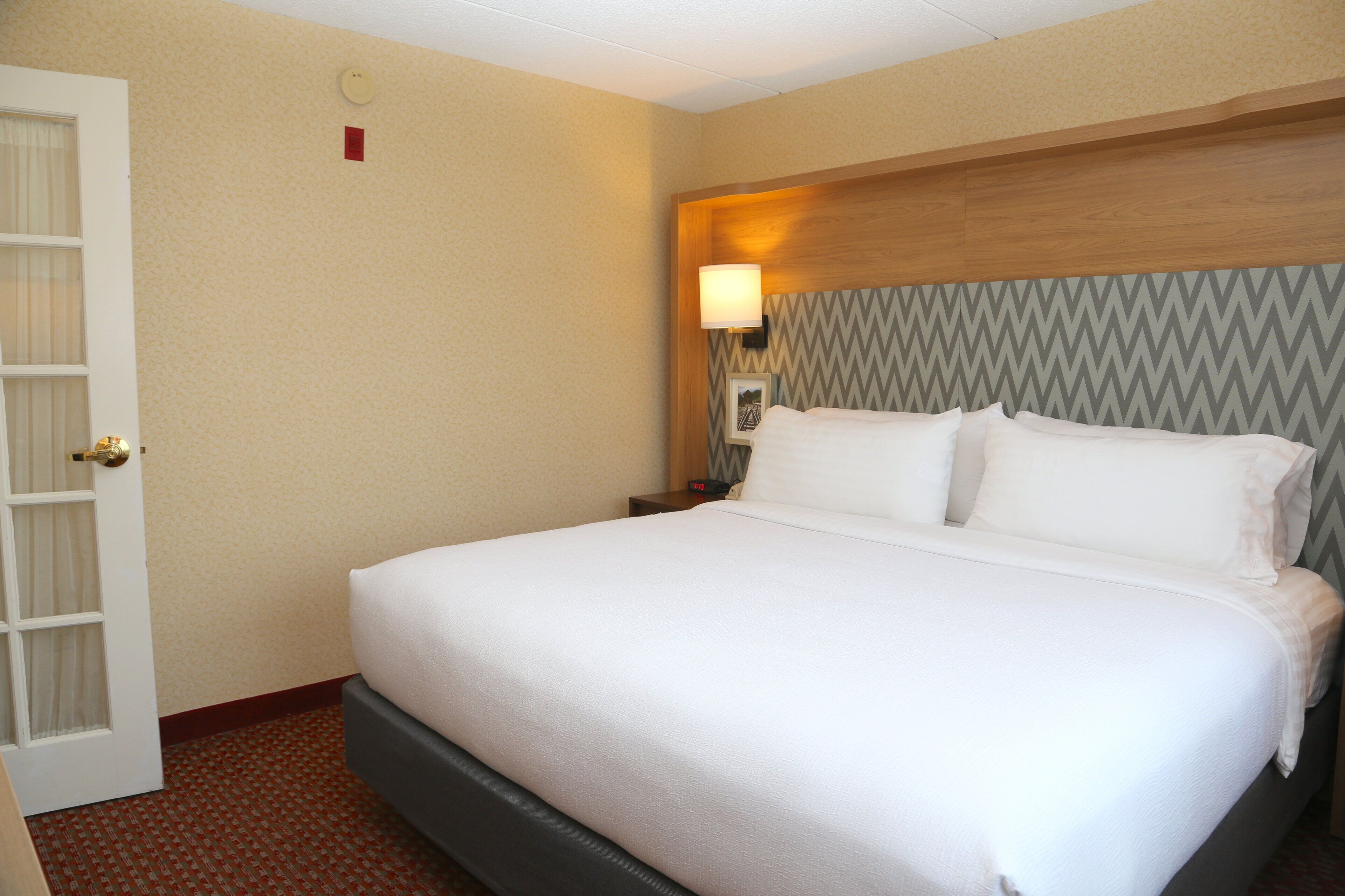 A hotel room with a bed, a nightstand, a lamp, and a door.