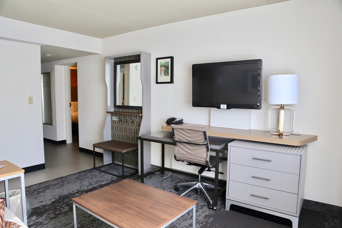 A hotel room with a desk, chair, TV, lamp, and a bed.