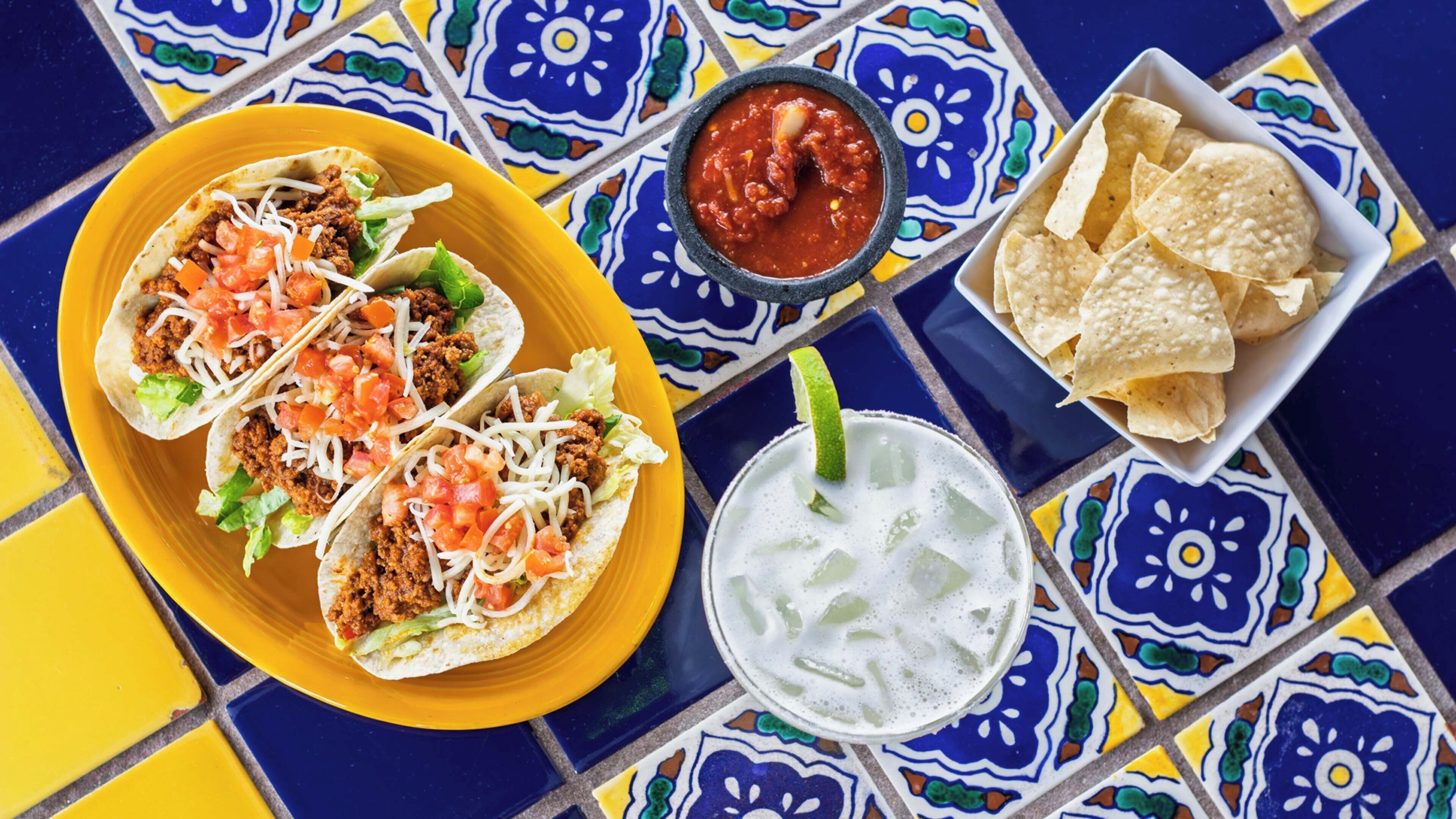 A table with tacos, a bowl of salsa, chips, and a drink.