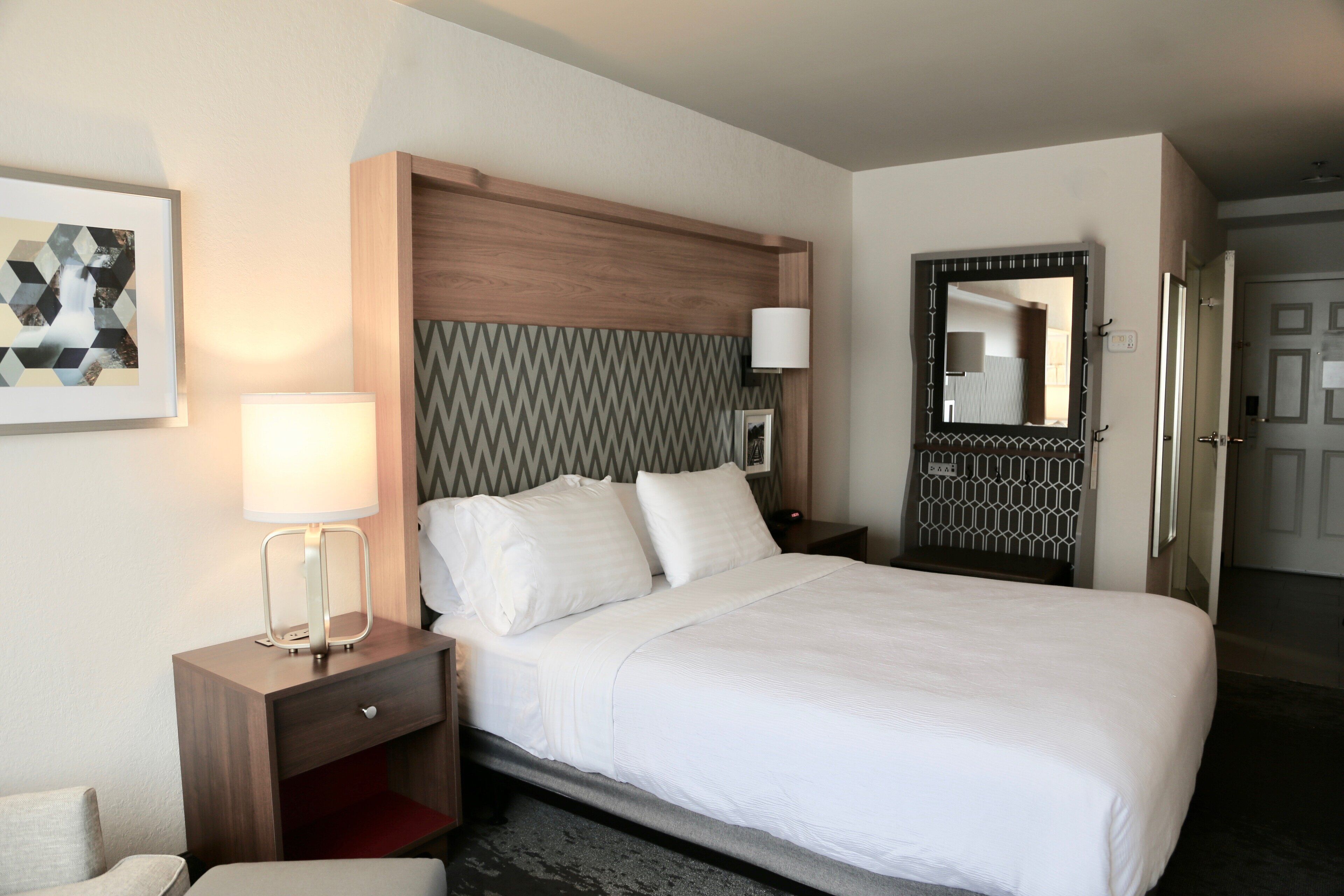 A hotel room with a bed, a nightstand with a lamp, a wall-mounted mirror, and a framed picture.