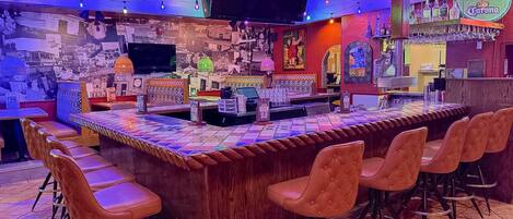 A bar area with a wooden counter, cushioned bar stools, and a variety of bottles and glasses on the shelves.