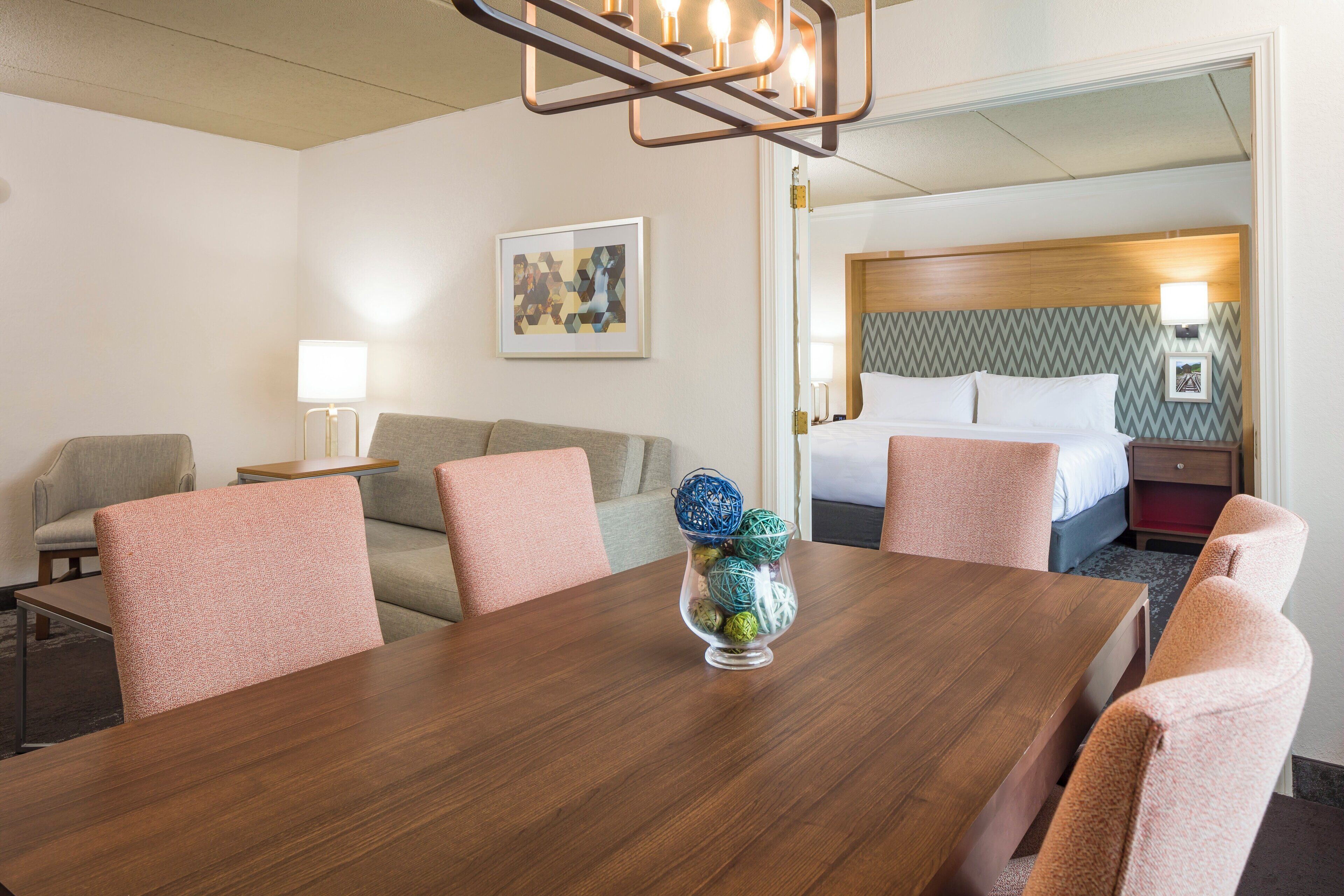 A hotel room with a wooden dining table, a sofa, a bed with a headboard, and a wall-mounted lamp.