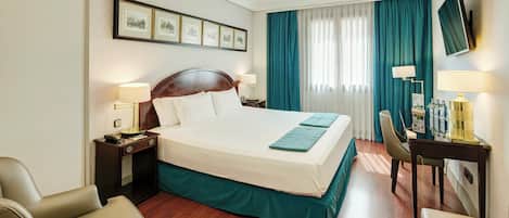 A hotel room with a large bed, two bedside tables with lamps, a desk with a chair, a TV mounted on the wall, and a window with curtains.
