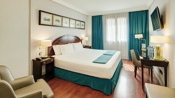 Una camera d'hotel con un letto grande, due comodini con lampade, una scrivania con sedia, una TV montata a parete e una finestra con tende.