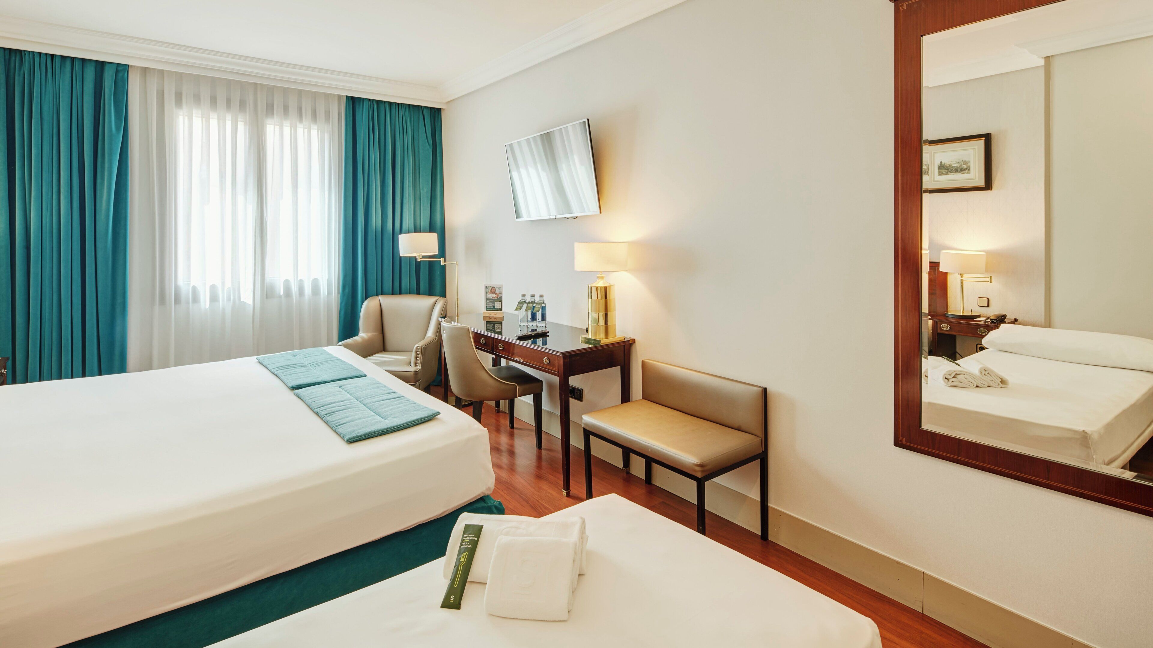 A hotel room with a bed, a desk, a chair, a mirror, and a window with curtains.