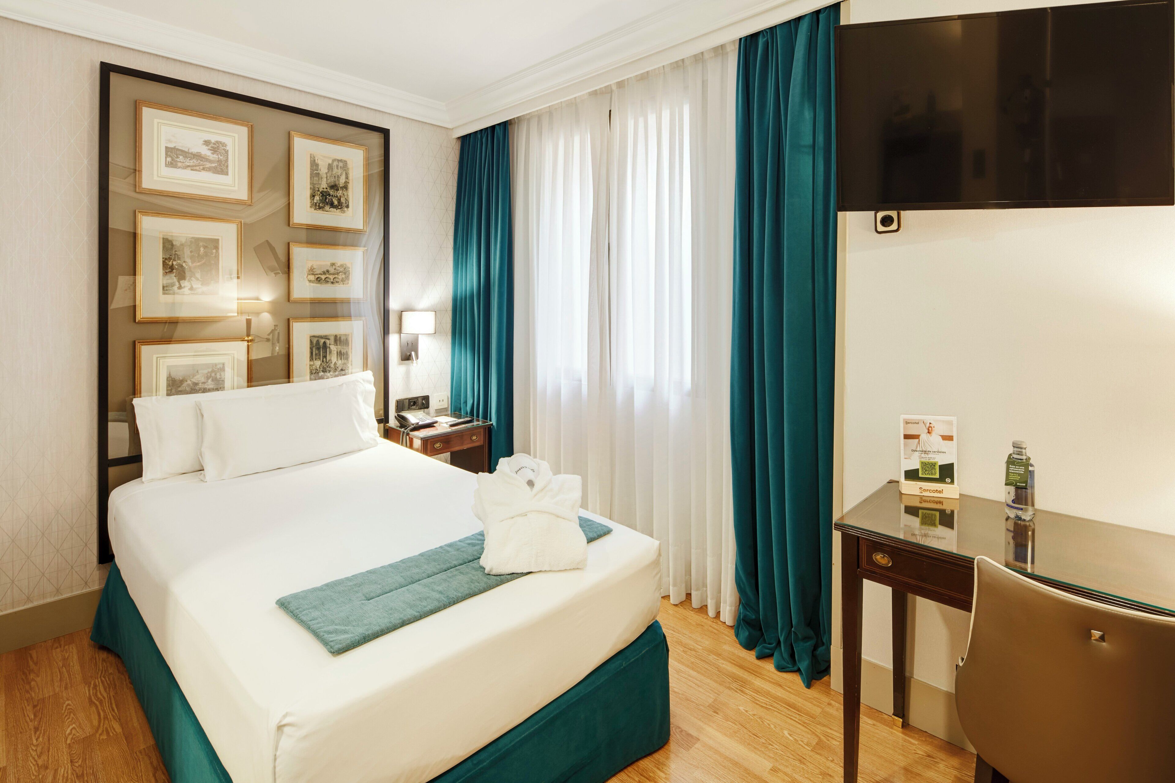 A hotel room with a bed, a desk, a TV, and framed pictures on the wall.
