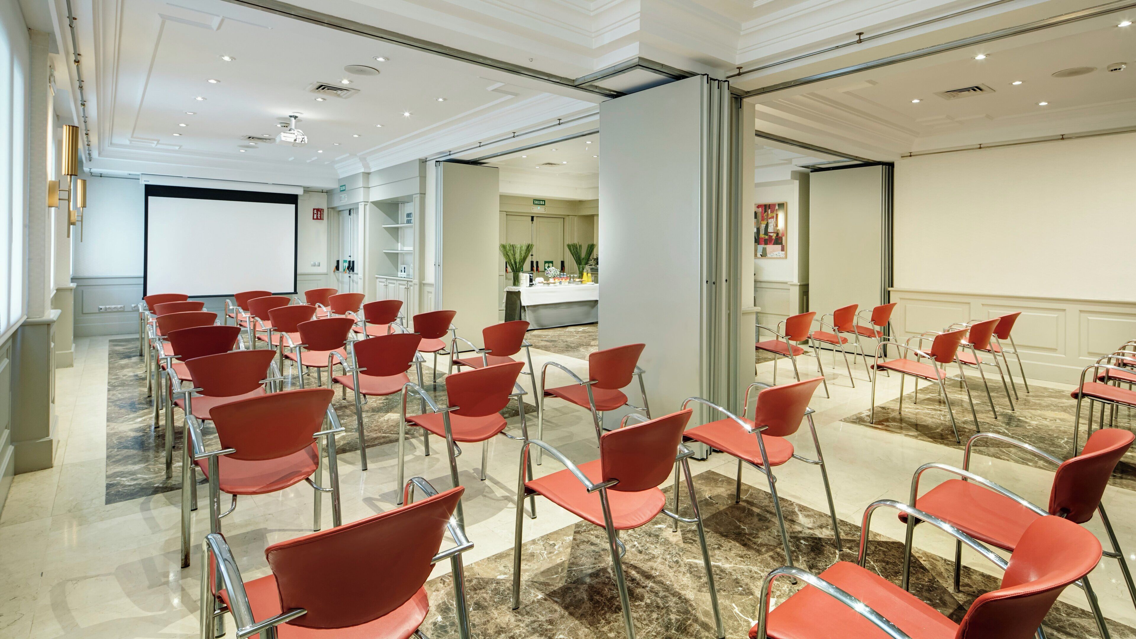A conference room with rows of red chairs, a projector screen, and a reception area in the background.