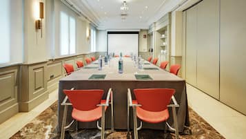 Een vergaderzaal met een lange tafel, rode stoelen, waterflessen en een projectorscherm aan het einde.