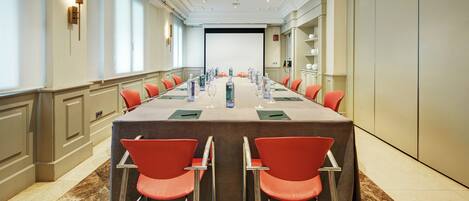 A conference room with a long table, red chairs, water bottles, and a projector screen at the end.