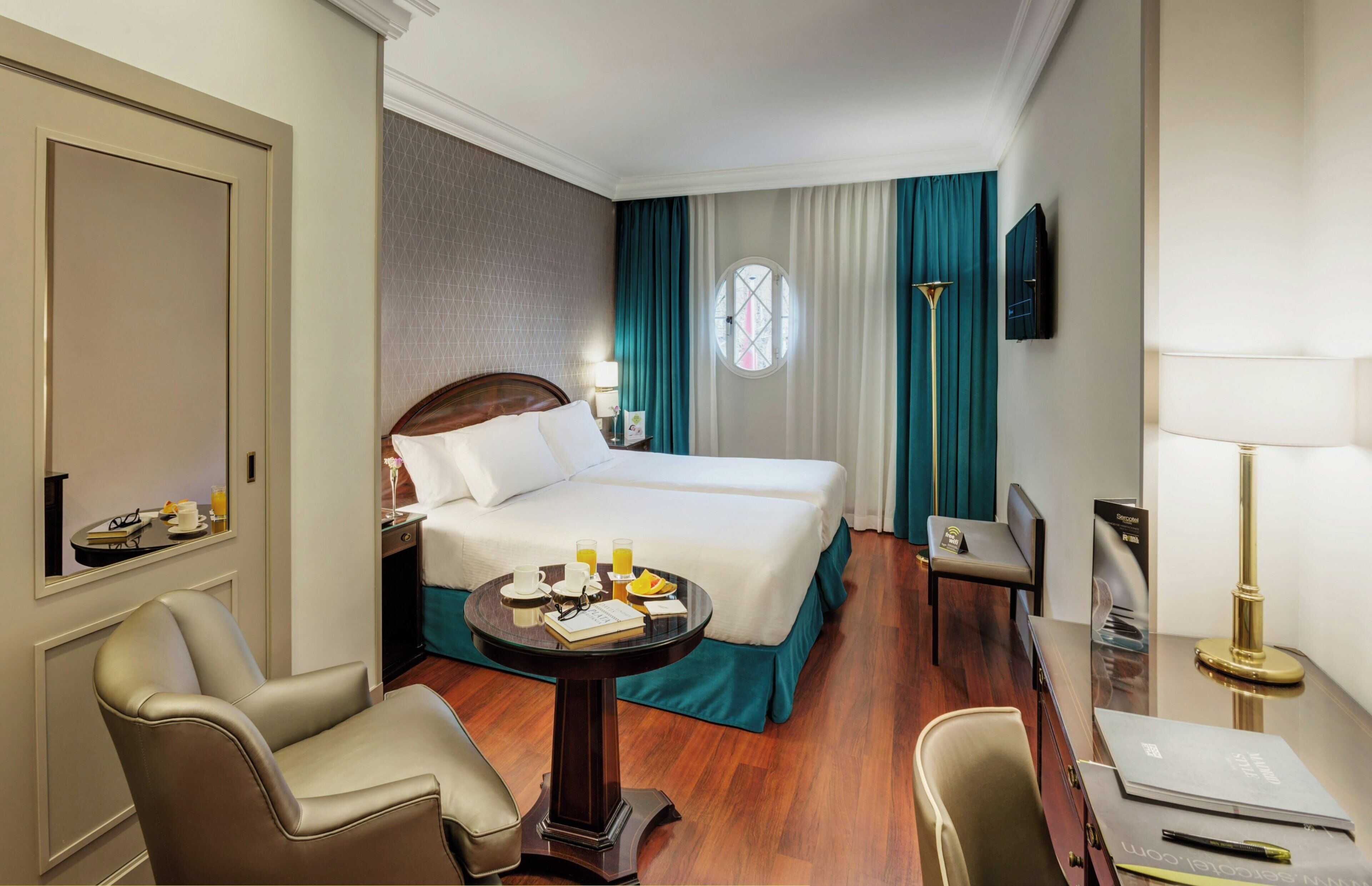 A hotel room with a bed, a desk, a chair, a table with a lamp, and a TV on the wall.