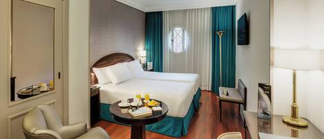 A hotel room with a bed, a desk, a chair, a table with a lamp, and a TV on the wall.