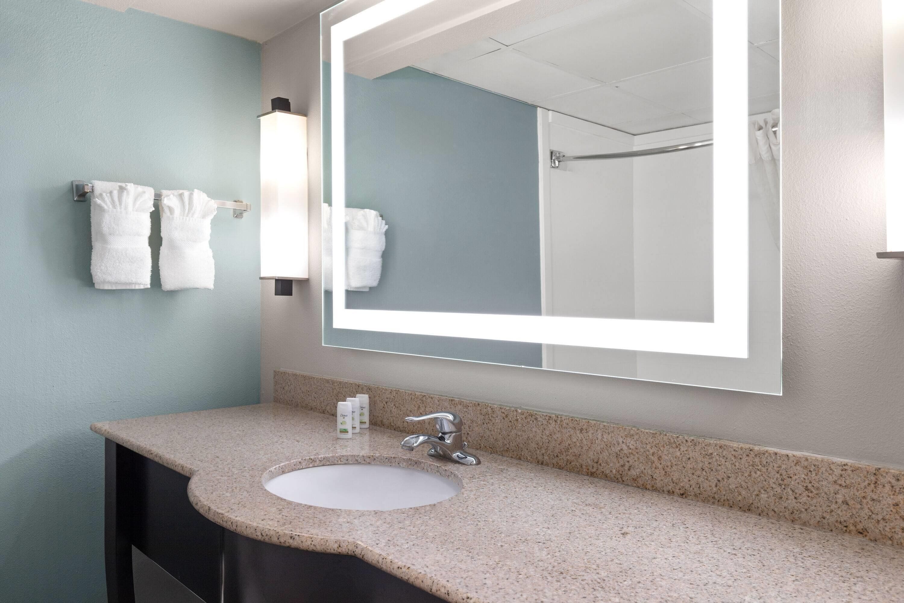 Une salle de bain avec un lavabo doté d’un plan de travail en granit, un grand miroir et deux serviettes suspendues à un porte-serviettes.
