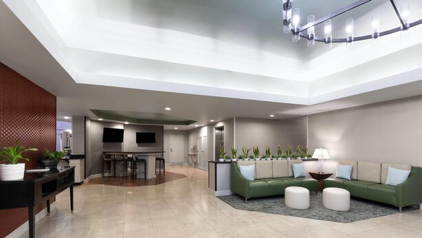 A spacious living area with a green sofa, a coffee table, and a dining table with chairs. There is a kitchen area in the background with a TV and a refrigerator. - Wingate by Wyndham Greensboro/Coliseum (Greensboro)