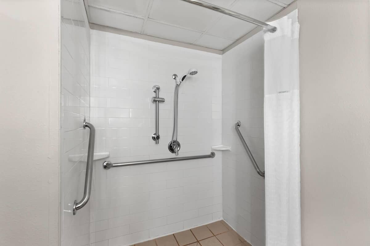 Un espace douche avec des murs recouverts de carreaux blancs, une barre d’appui et un pommeau de douche.