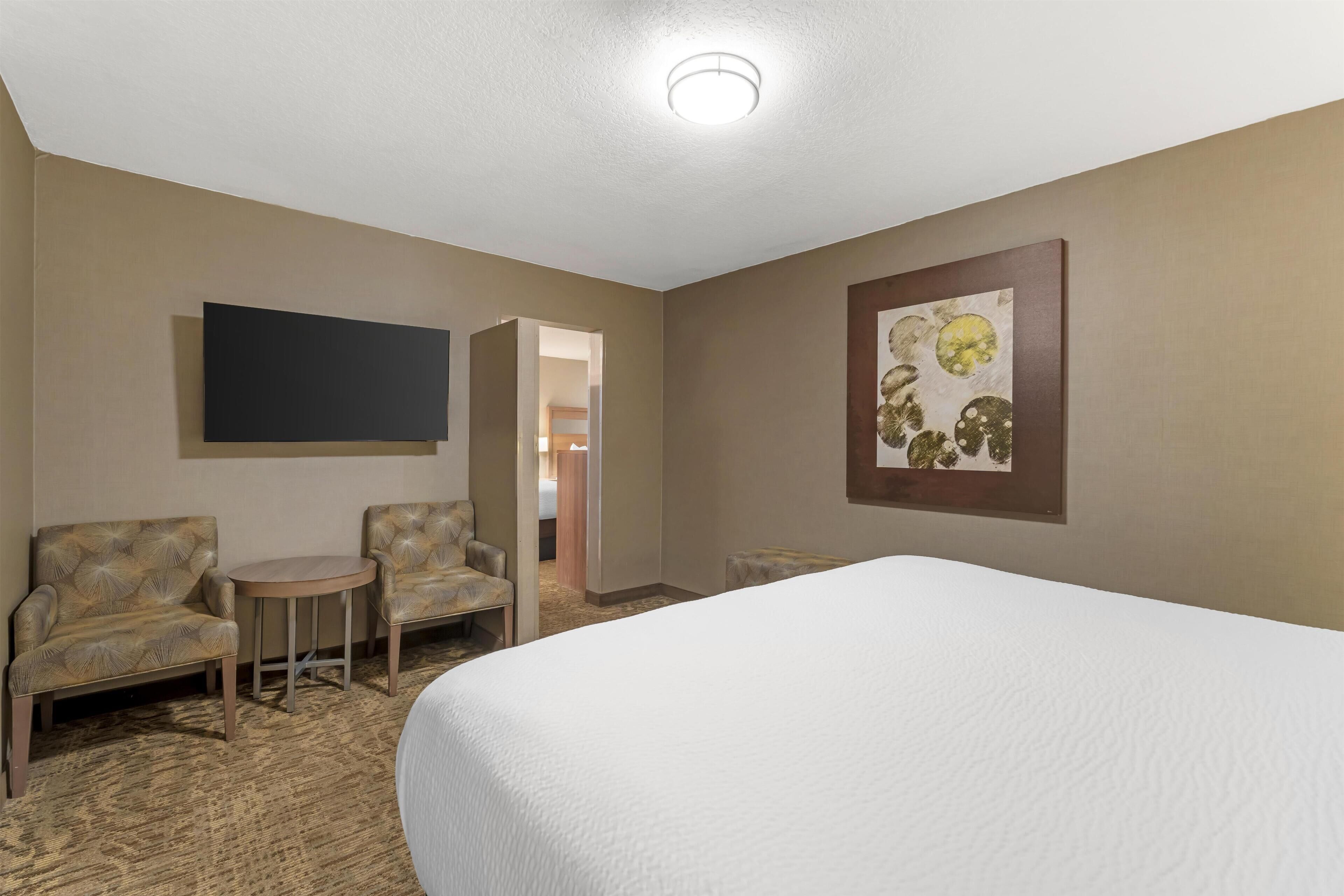 A hotel room with a bed, two armchairs, a small table, a flat-screen TV mounted on the wall, and a framed artwork above the bed.