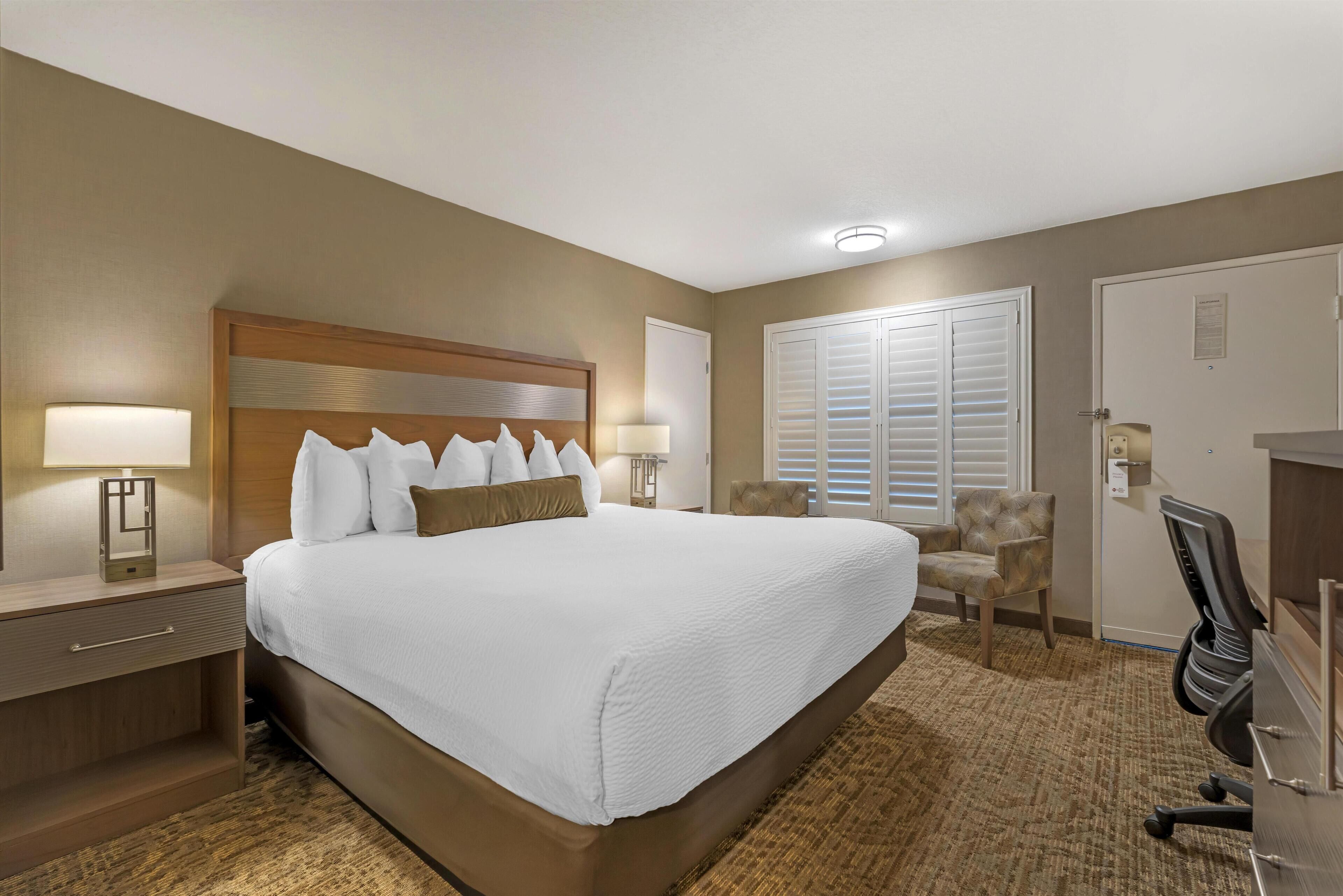 A hotel room with a large bed, two bedside lamps, a chair, a desk, and a window with blinds.
