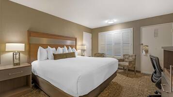 A hotel room with a large bed, two bedside lamps, a chair, a desk, and a window with blinds.