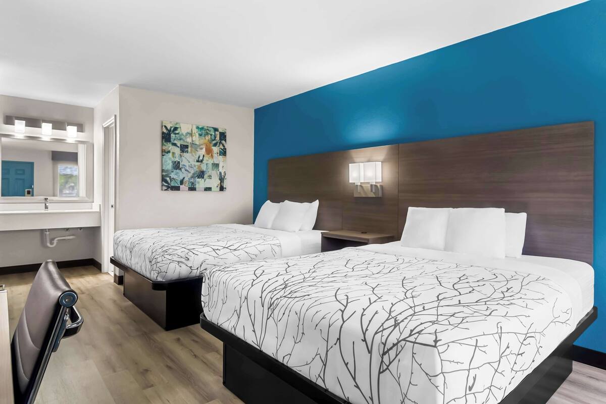 A hotel room with two beds, a large window, a bathroom with a mirror, and a blue accent wall.