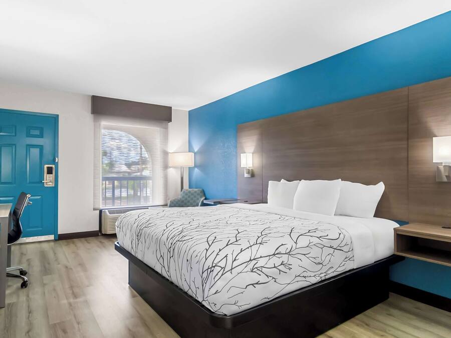 A modern hotel room with a large bed, a desk, and a chair. There is a window with a view of the outdoors.