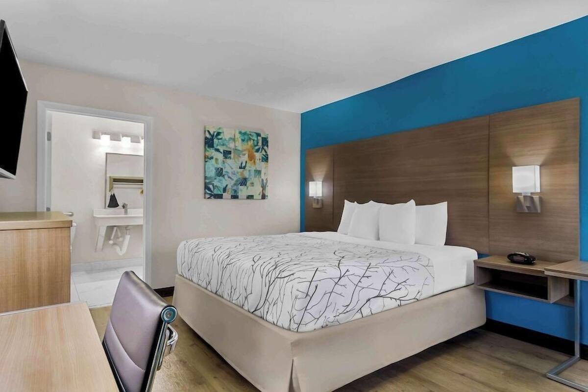 A hotel room with a bed, bedside table, a mirror, a painting, and a TV.