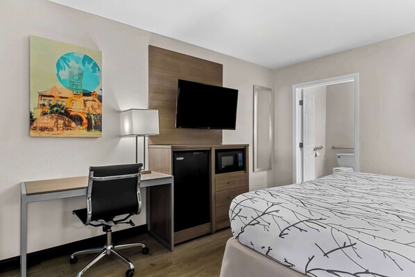 A hotel room with a bed, desk, chair, television, microwave, and a painting on the wall.
