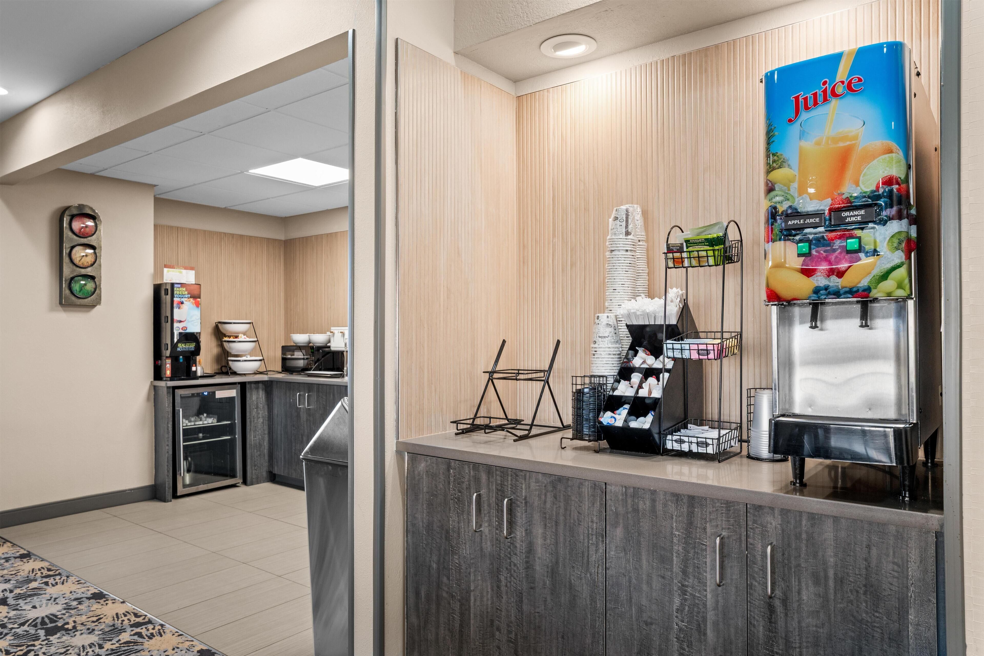 A hotel reception area with a juice dispenser, coffee machine, and a small refrigerator.